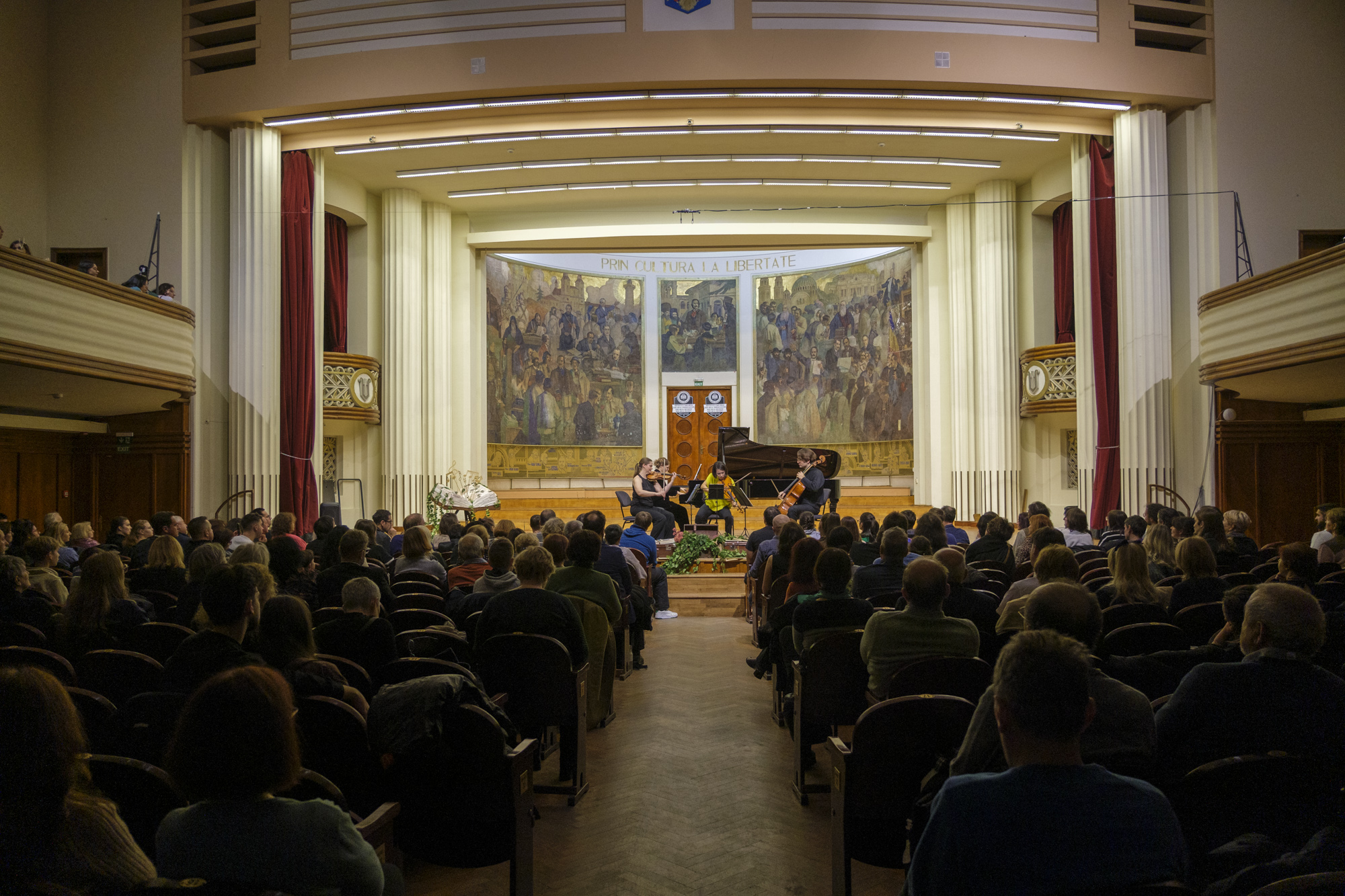 A string quartet performs on stage in front of a mural in a concert hall, with an audience seated in the foreground, concert sala Auditorium UBB/ Sala Auditorium Maximum